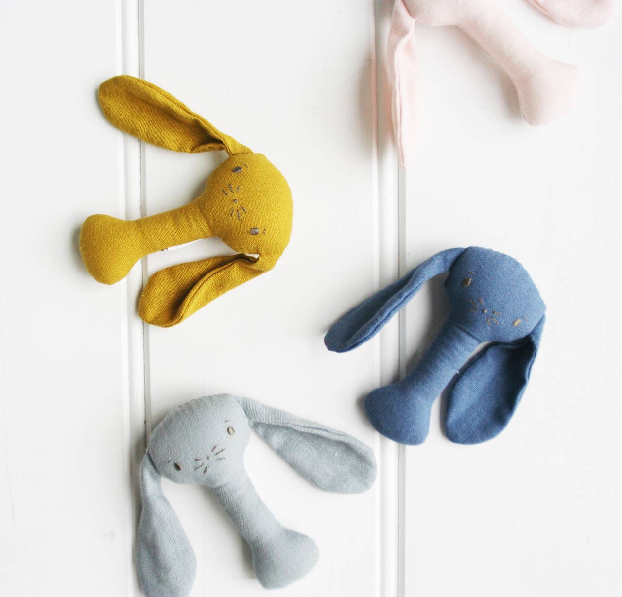 Heirloom-style bunny stick rattles in grey, mustard, blush and blue linen, photographed from above.