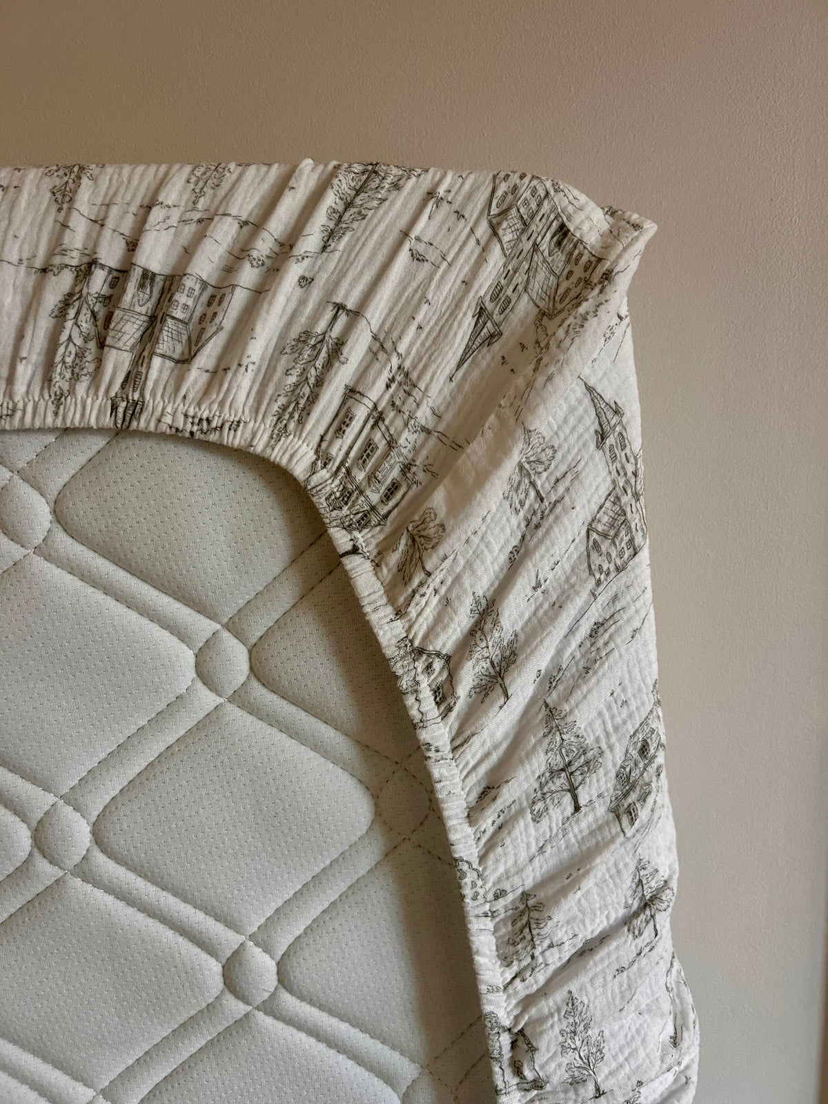 A close-up of a fitted sheet with a sage green toile print on a white background, draped over a white quilted mattress.